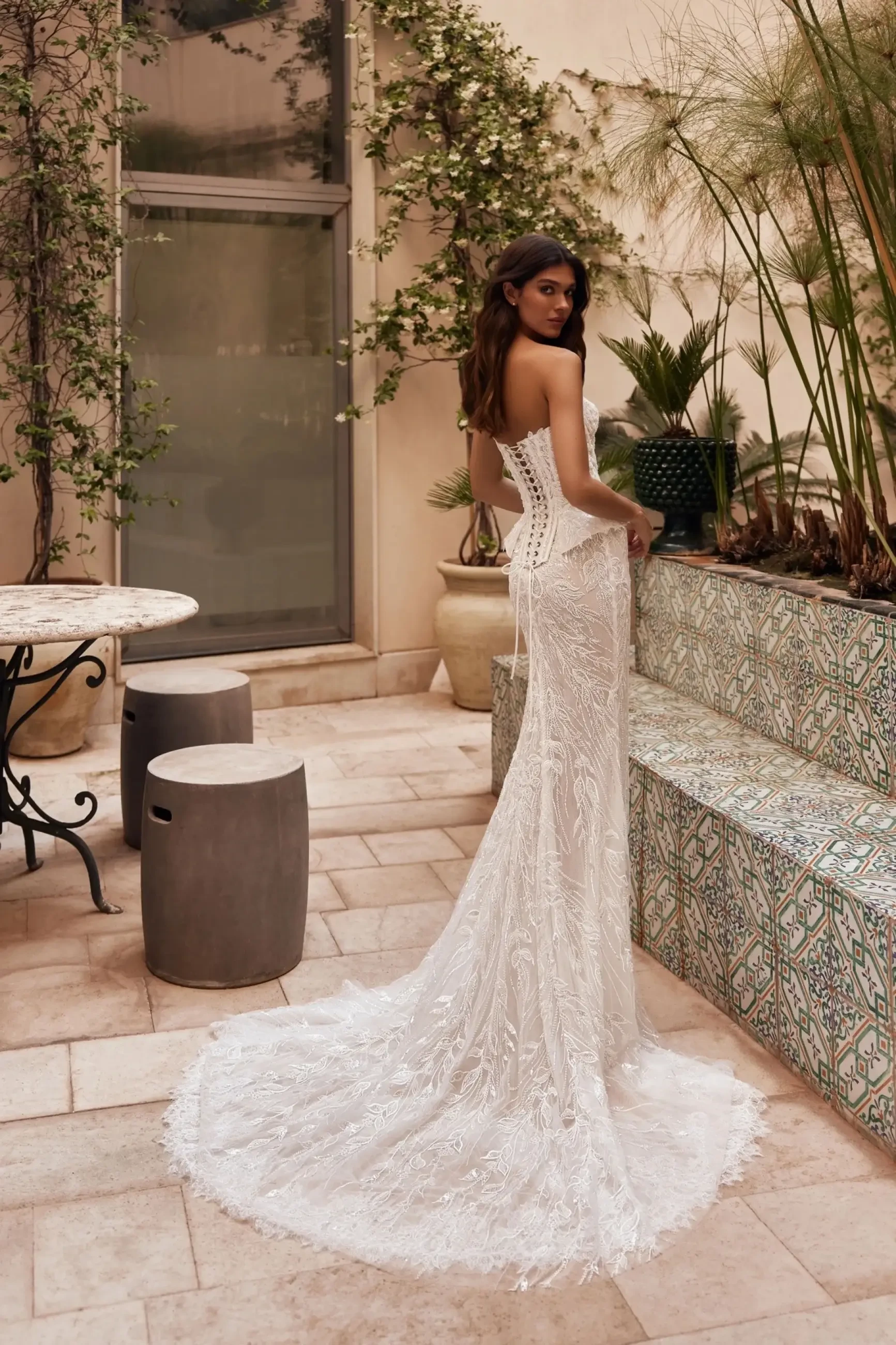 A woman in a stunning wedding dress stands with her back to the camera, facing a beautifully landscaped area with plants and decorative tiles. The dress features intricate lace details and a long train, while the setting includes stone tables and modern seating.