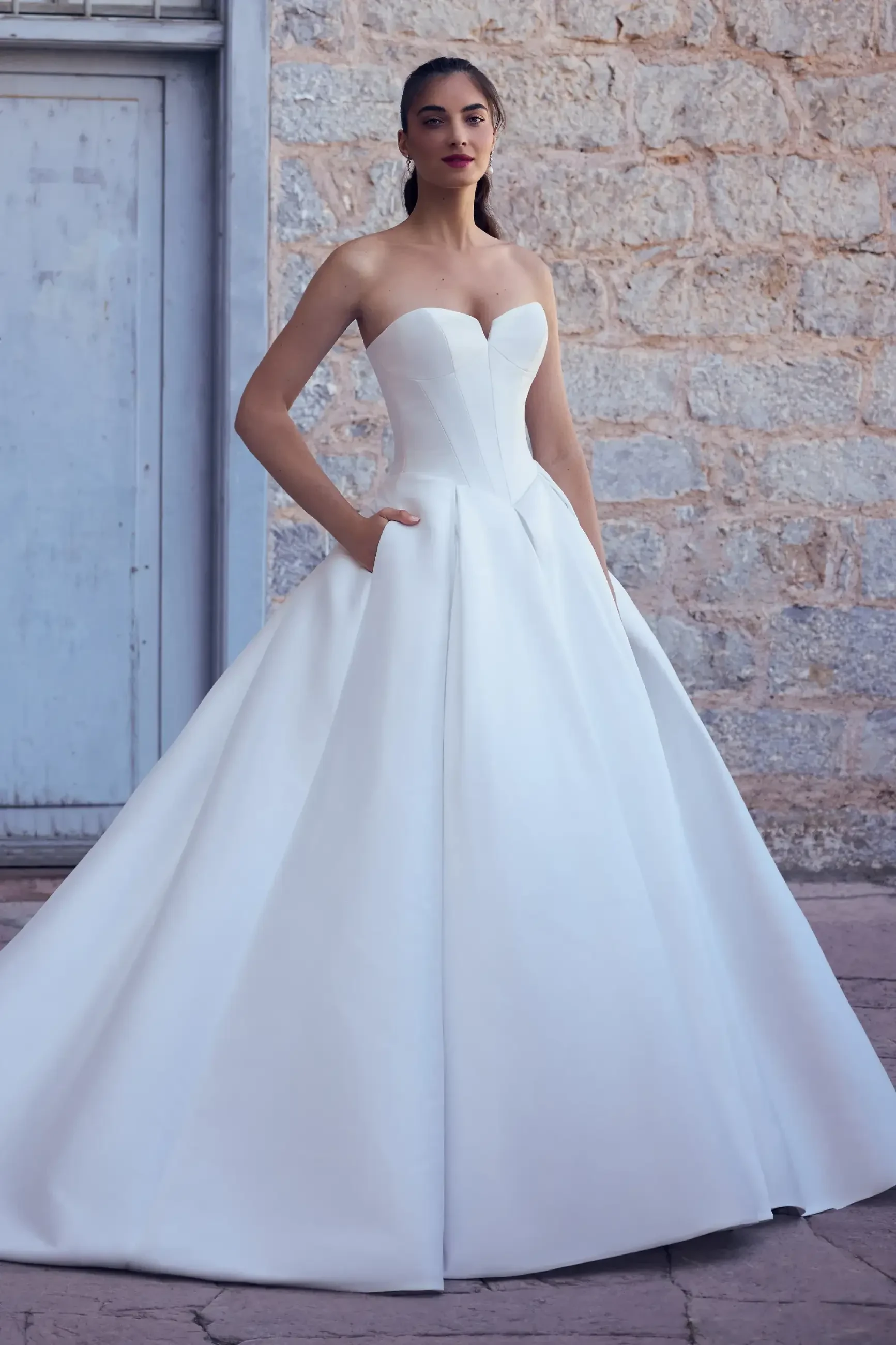 A woman in a strapless white ball gown stands confidently against a rustic stone wall. Her hands are in the dress pockets, exuding elegance and modern style.