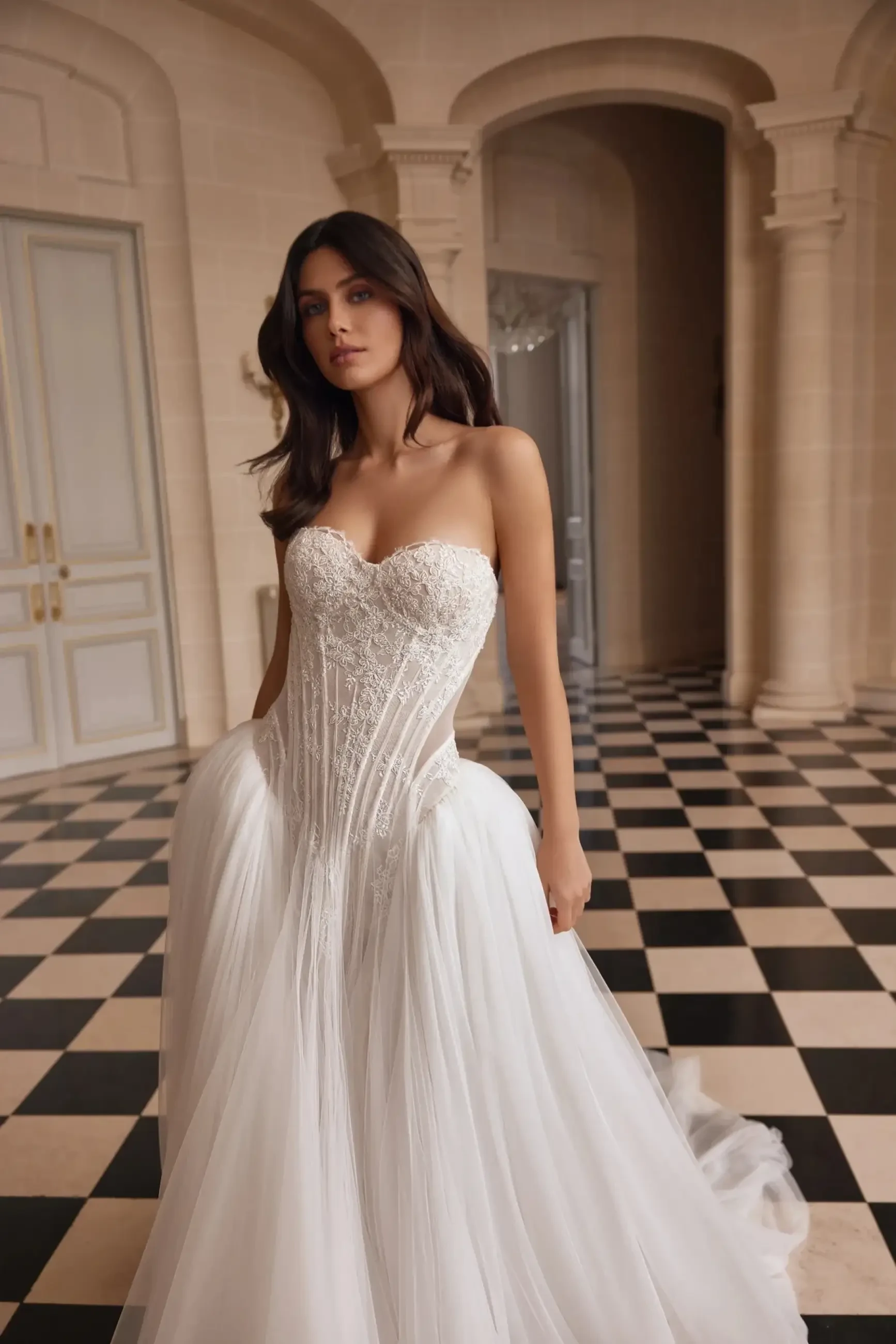 A woman in a white strapless wedding dress with intricate detailing, standing in an elegant interior with a checkered floor.