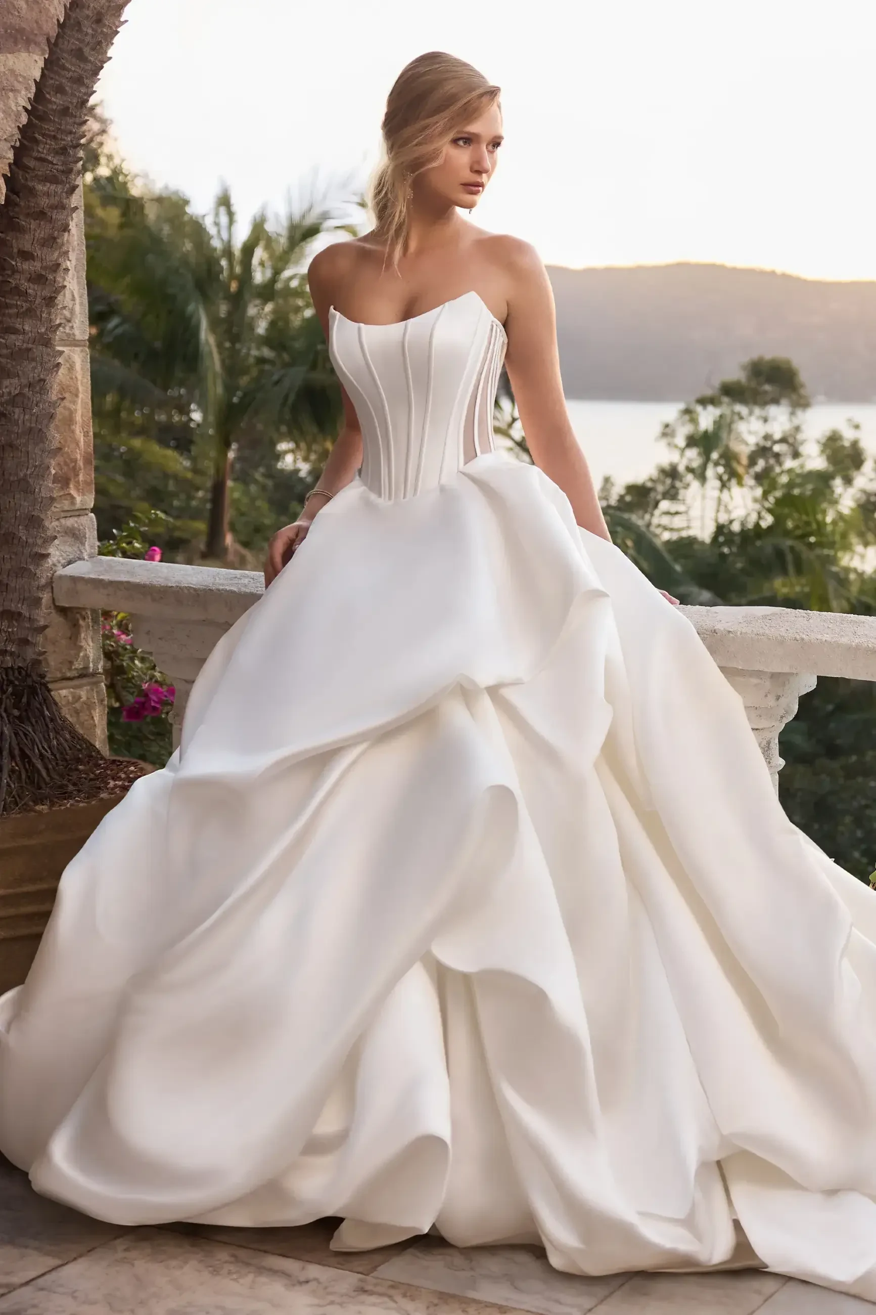 Woman in an elegant strapless white wedding gown with a voluminous skirt stands on a terrace, surrounded by lush greenery at sunset.