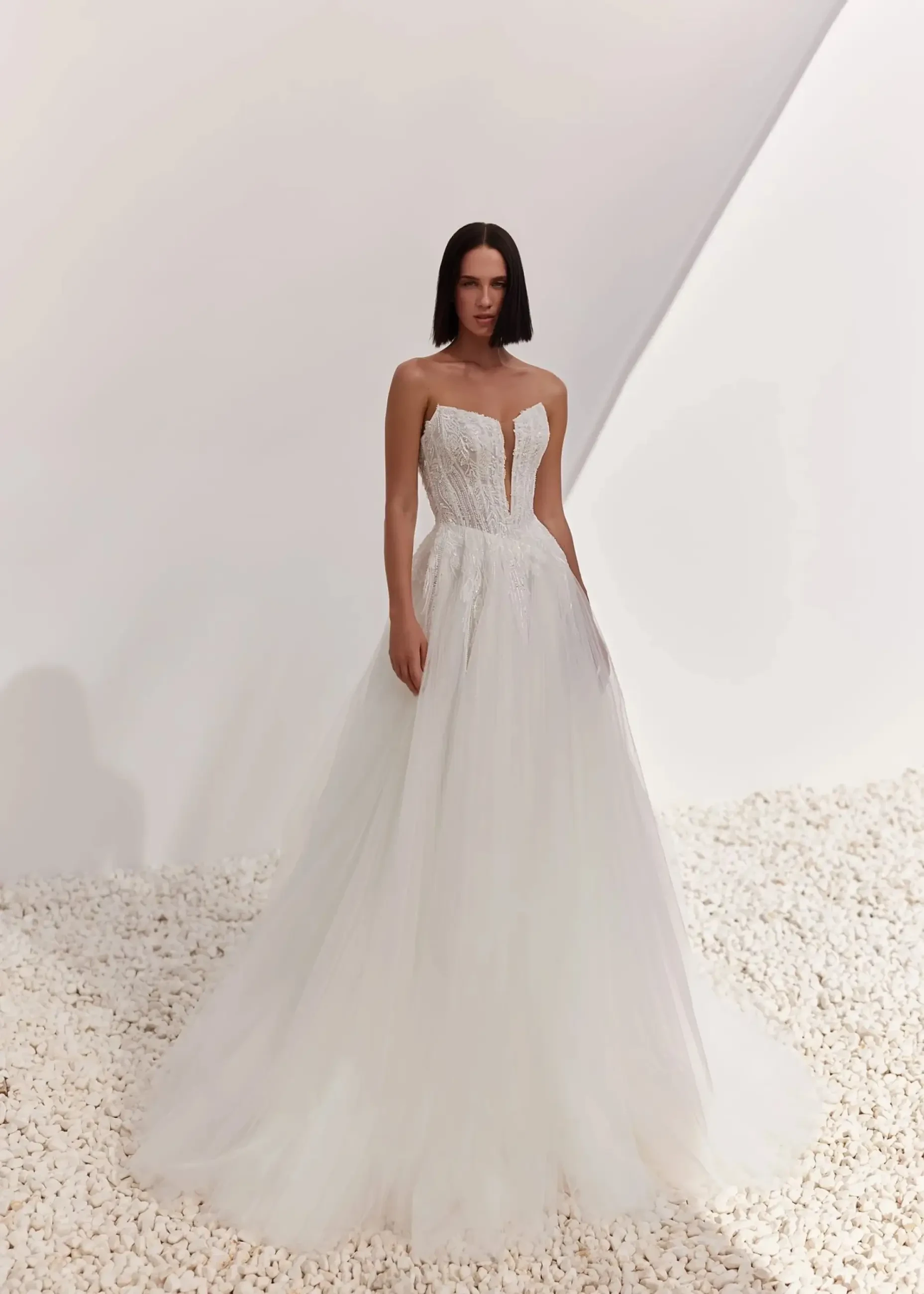 A model wearing a strapless white wedding gown with a fitted bodice and flowing skirt, standing against a minimalist background.