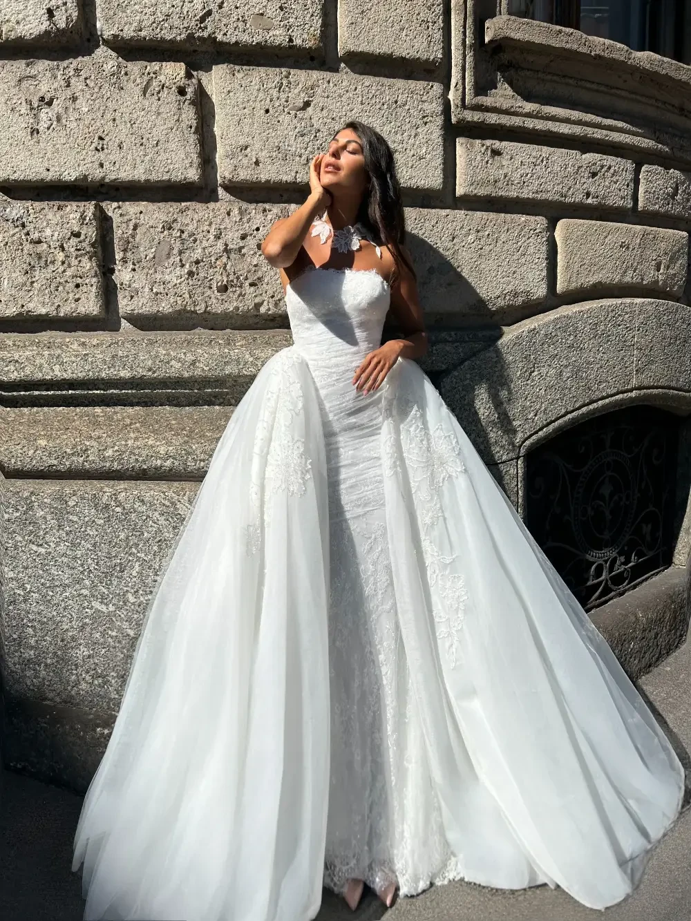 A woman in a white dress with a long train poses against a stone wall.