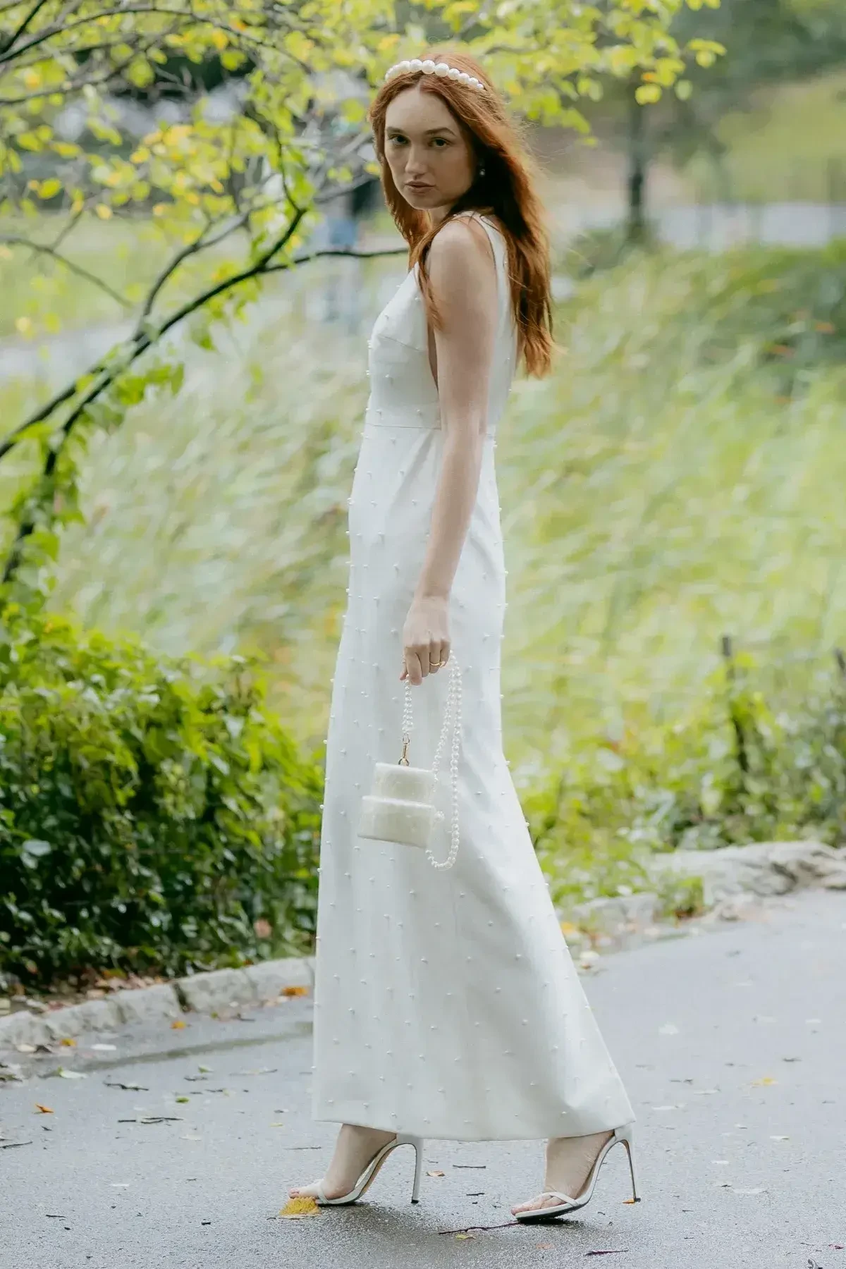 A woman in a white dress and headband stands outdoors, holding a small handbag, with greenery in the background.