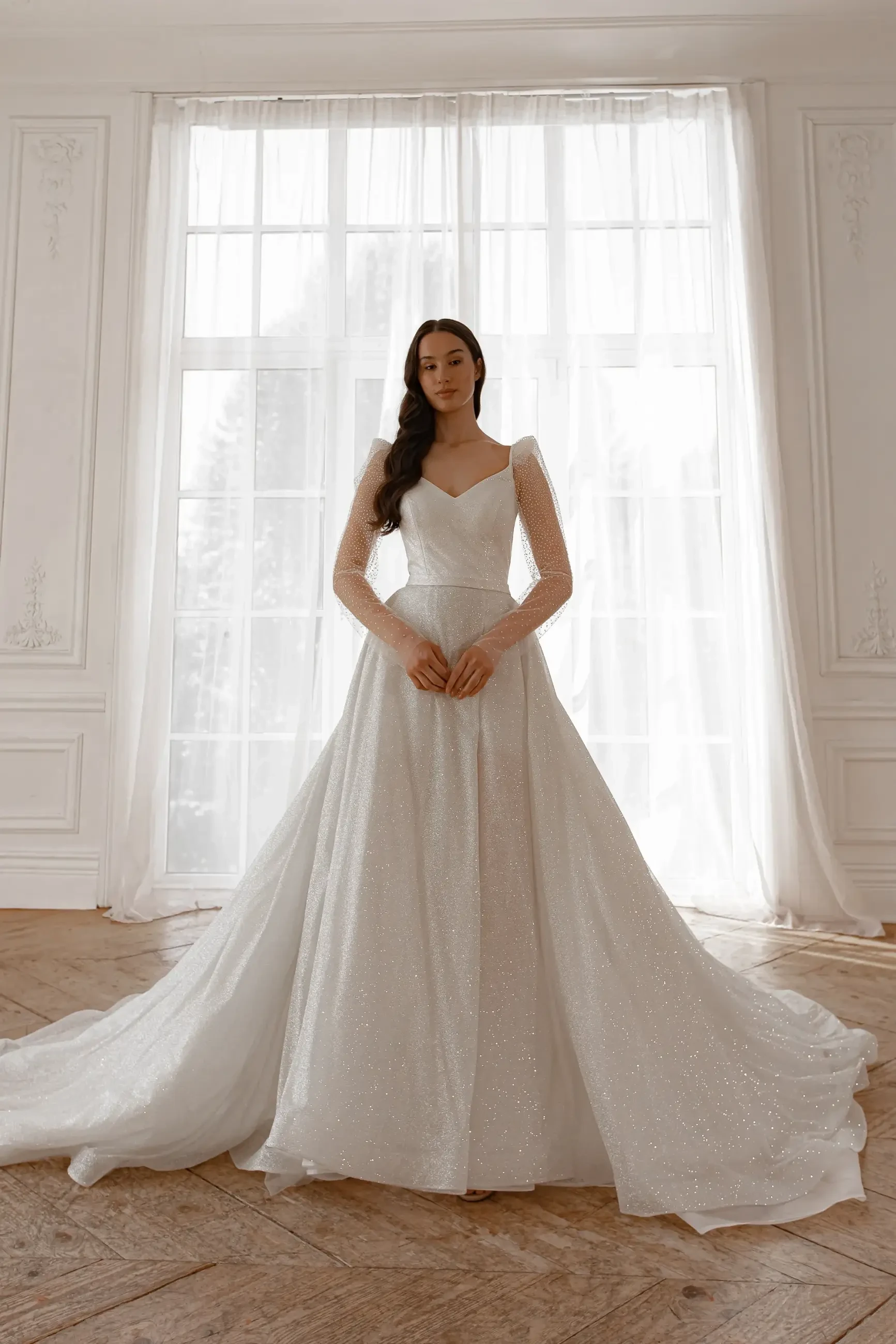 Woman in a sparkling white wedding gown with long sleeves stands gracefully in a bright room. Sheer curtains and elegant decor create a serene atmosphere.
