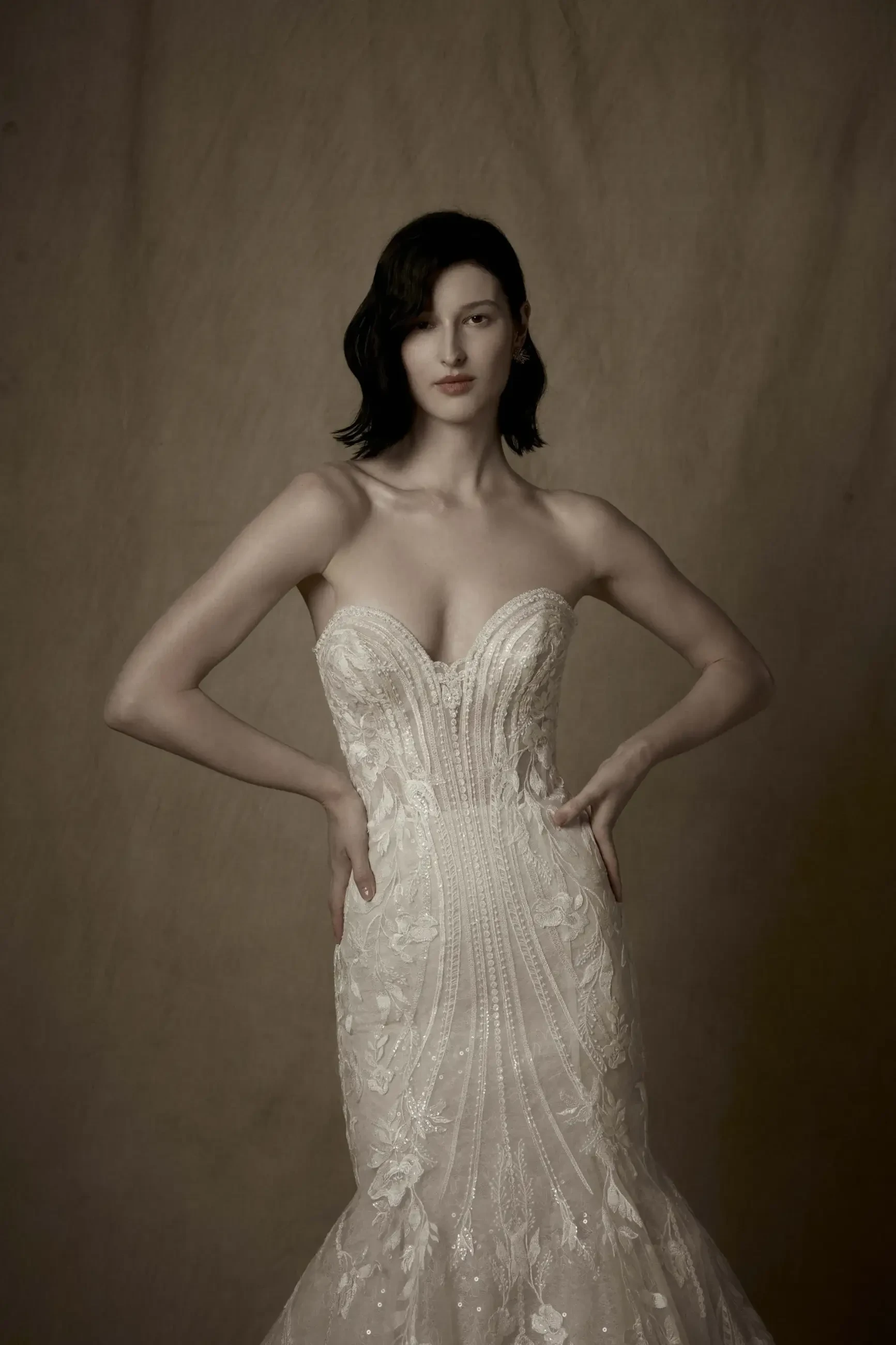 A woman wearing an elegant, fitted wedding dress with intricate beading and floral designs, standing with her hands on her hips against a muted background.