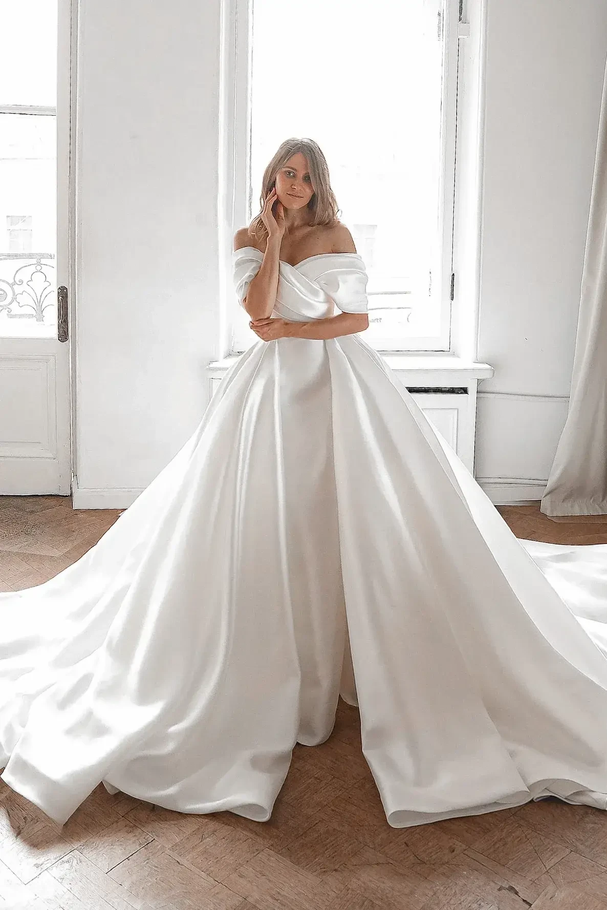 Woman in elegant off-the-shoulder white satin wedding gown stands in a bright room with wooden floors and large window, projecting grace and poise.