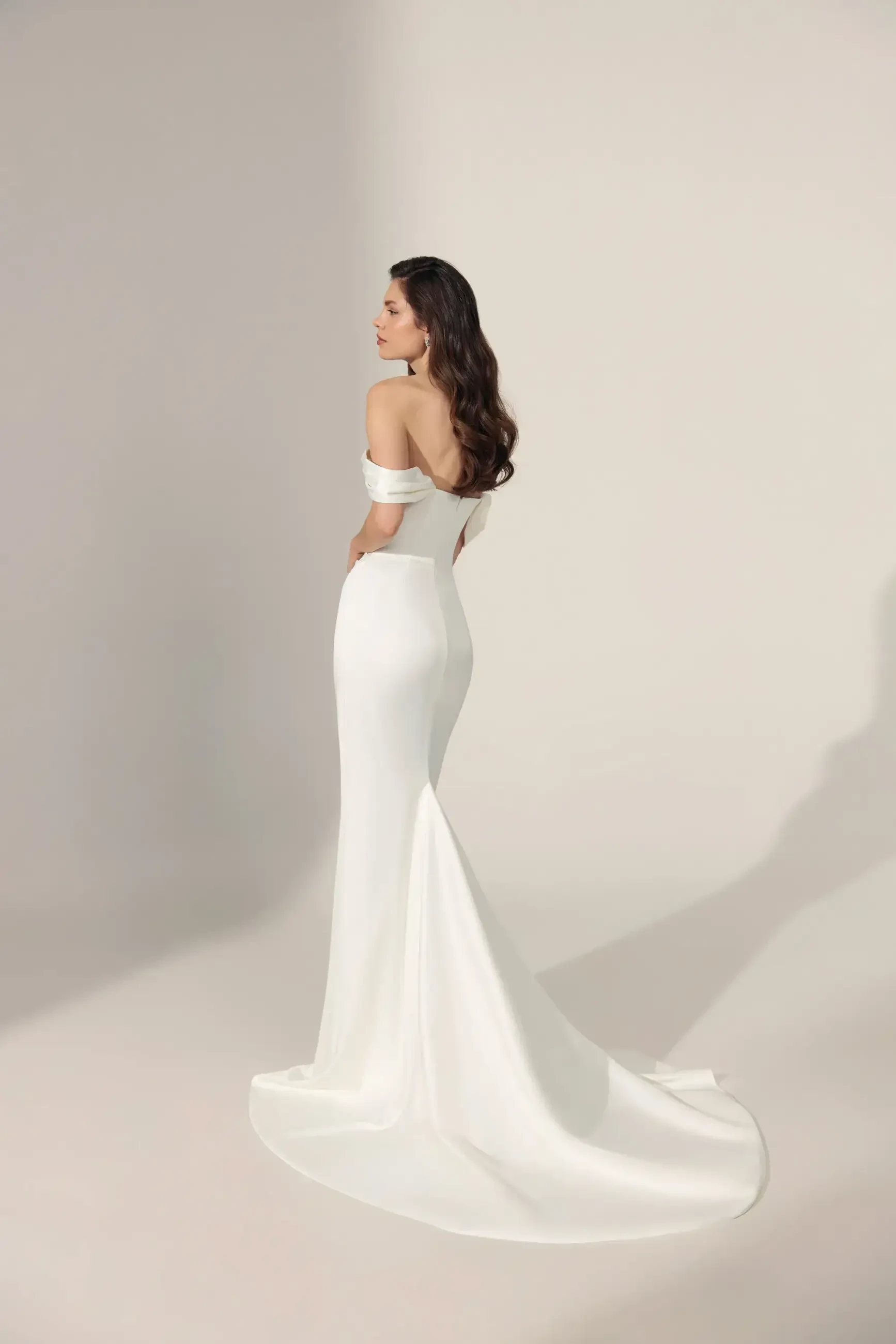 A woman in an elegant, off-the-shoulder white gown with a long train stands in profile against a neutral background. Her expression is serene and contemplative.