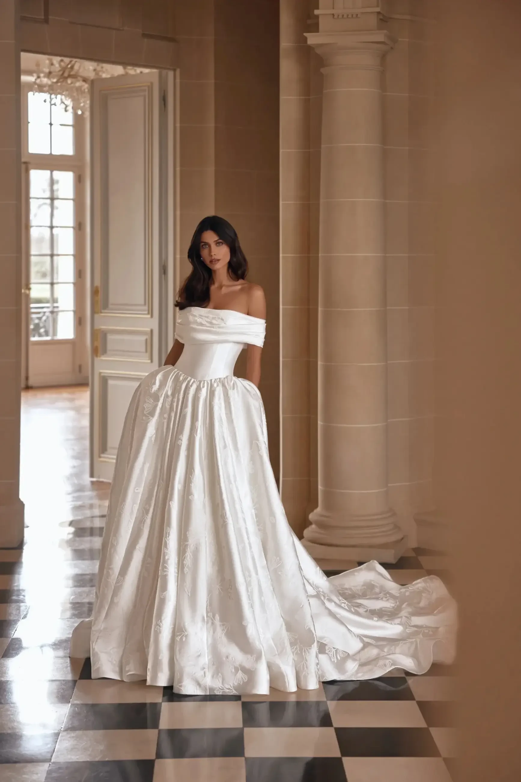 A woman in a white off-the-shoulder gown stands in an elegant interior with checkered flooring and large windows.