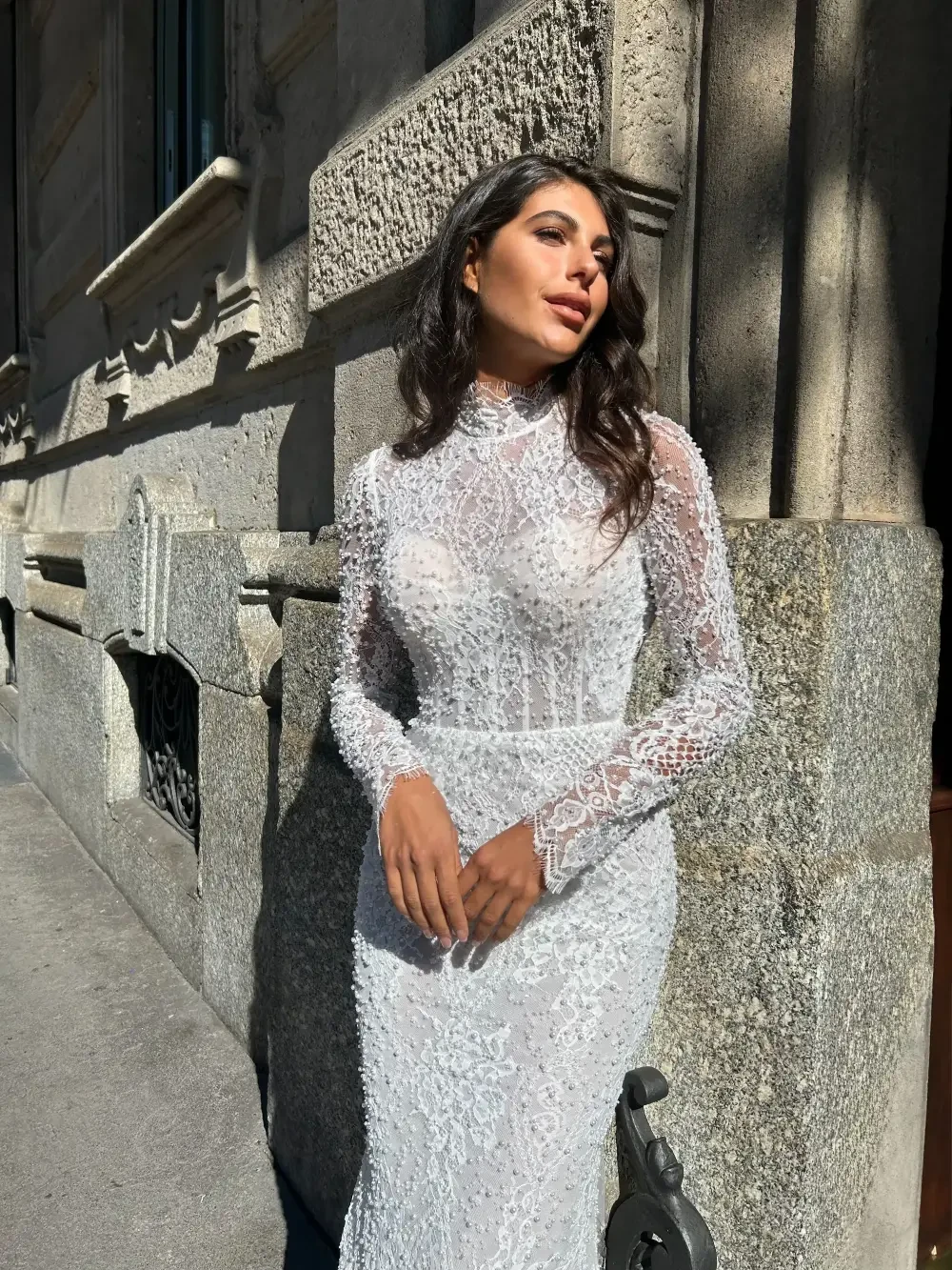 A woman in a sheer, lace white dress stands on a city street, bathed in sunlight, with a historical building in the background.
