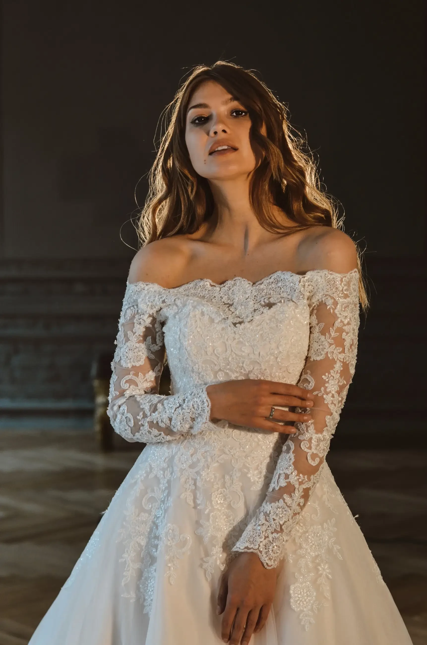 A woman in an elegant off-the-shoulder wedding dress with long lace sleeves stands gracefully, looking at the camera. The background is softly lit, enhancing the romantic atmosphere.