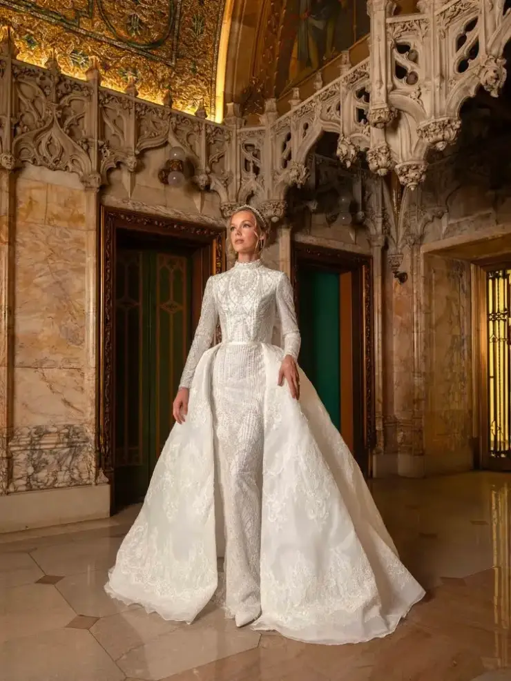 A model wearing an elegant white gown stands in an ornate architectural space with intricate details and gold accents.