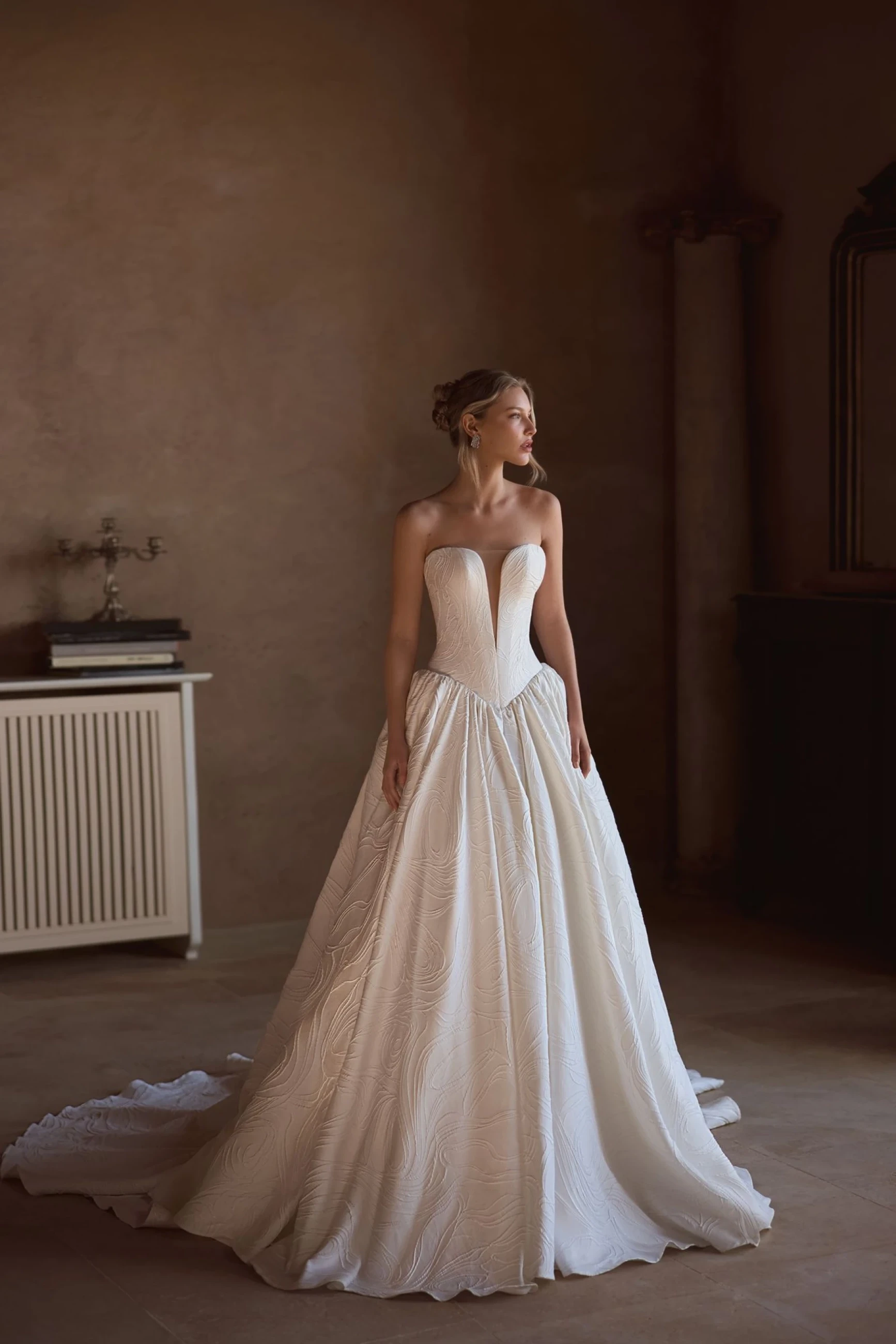 A model wearing an elegant wedding dress with a high slit, featuring intricate lace detailing, stands in a garden setting with potted plants and stone architecture.