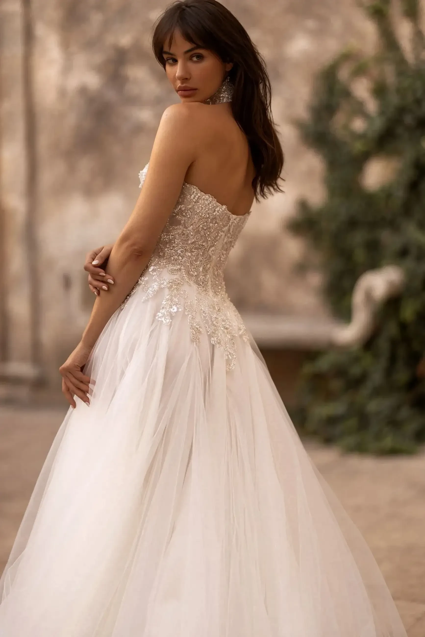 A woman in a strapless, embellished white gown stands with her back to the camera, looking over her shoulder. The background features a blurred, natural setting.