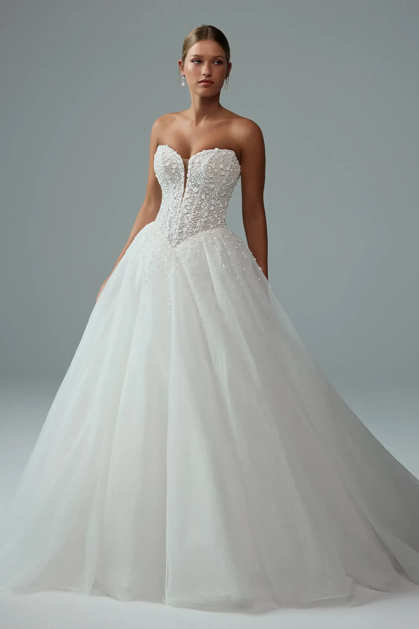 A model wearing a strapless wedding gown with an embellished bodice and a full tulle skirt, standing against a gray background.