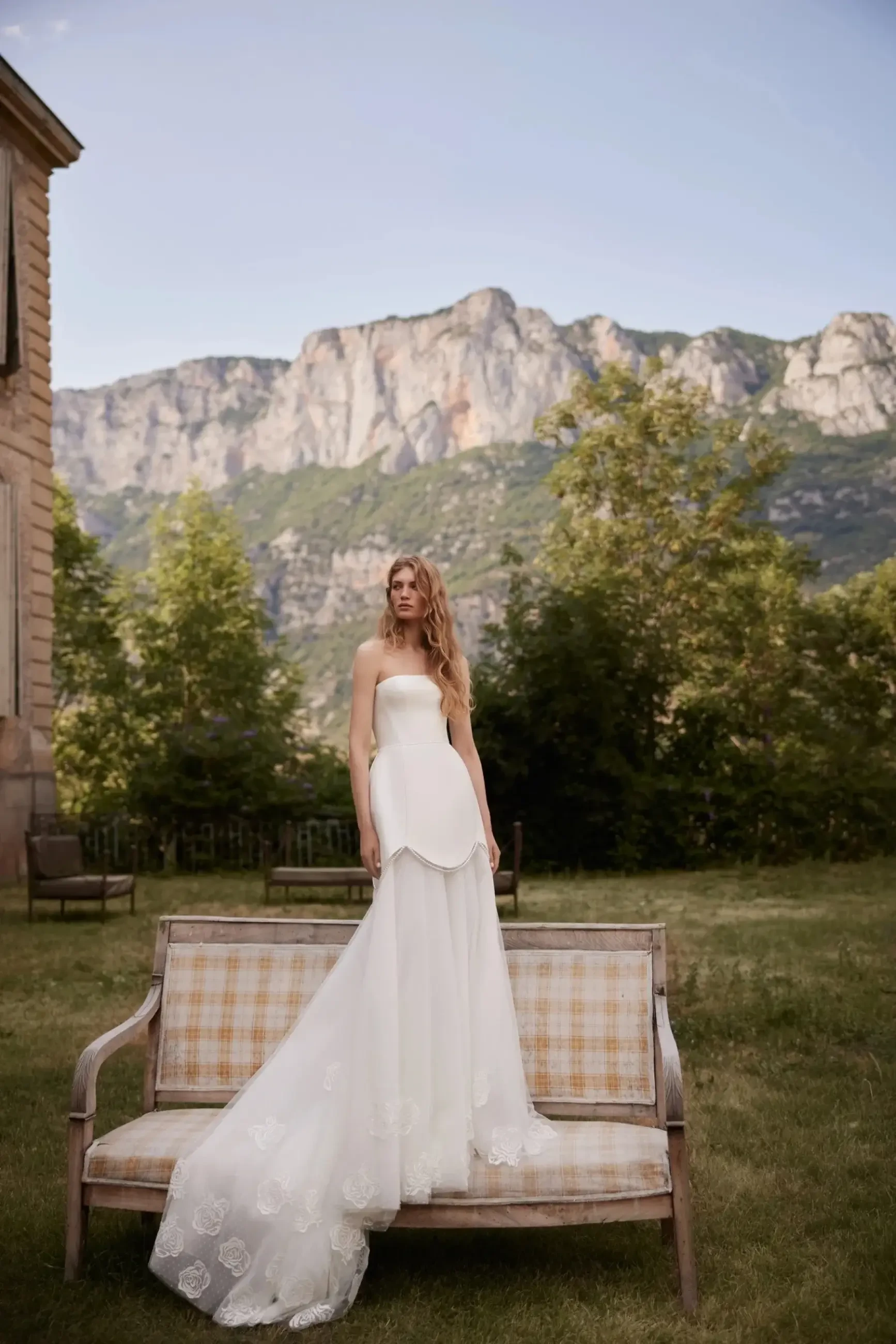 A woman in a strapless white wedding dress stands on a vintage sofa in an outdoor setting, with mountains and trees in the background.